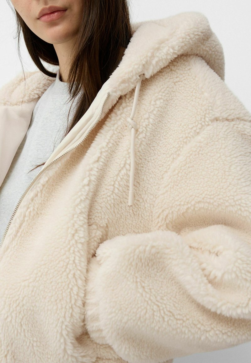 Veste à capuche en polaire crème avec une texture douce, dotée d'une fermeture éclair devant et de cordons de serrage sur la capuche.