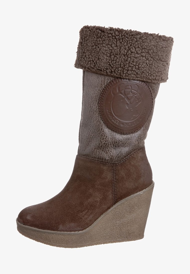 Brown wedge boot with a textured leather upper and a soft, fluffy brown cuff. Features a circular logo patch on the side.