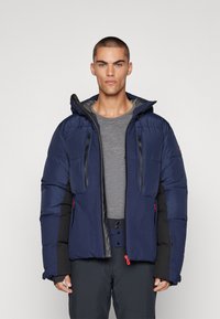 Navy blue puffy jacket with black accents, zippered pockets, and red zipper pull. Features a hood and textured fabric insulation.