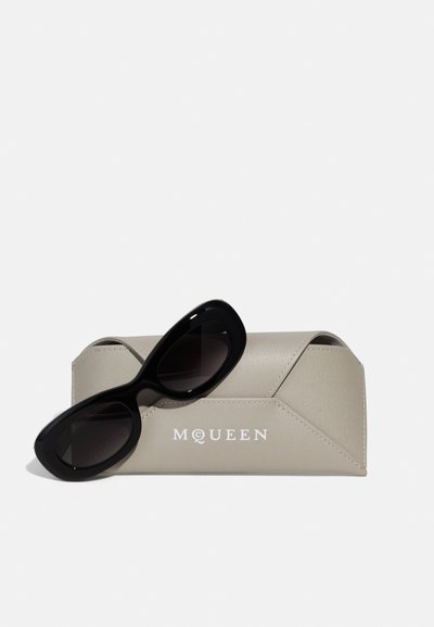 Fekete napszemüveg sima, lekerekített dizájnnal, egy világosszürke, textúrázott tokon pihen, amely geometriai mintázattal és a "McQueen" logóval díszített.