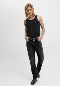 Joven con tatuajes que lleva una camiseta sin mangas negra, jeans negros, cinturón negro y botas negras, de pie con las manos en los bolsillos.