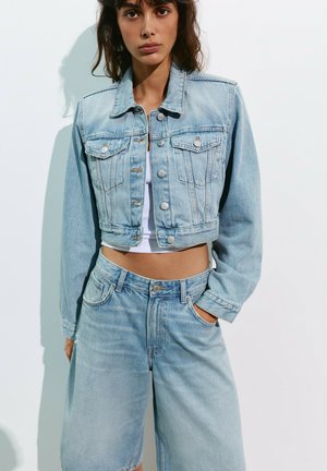 Femme portant une veste en denim courte bleu clair et un short en denim taille haute assorti, une main dans la poche, sur fond blanc.