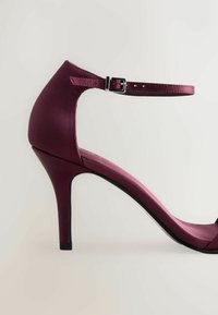 Zapato de tacón alto burdeos con acabado satinado, que presenta una correa en el tobillo con hebilla, un tacón de aguja delgado y un diseño de punta abierta.