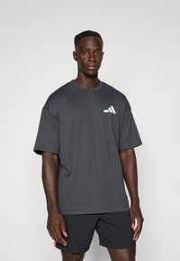 Mies, który ma na sobie ciemnoszary, oversize'owy T-shirt z małym białym logo na lewej piersi oraz czarne szorty sportowe z czerwonymi akcentami.
