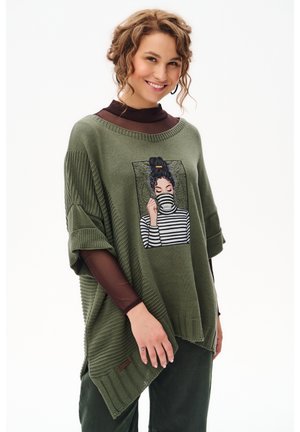 Kobieta o kręconych włosach ubrana w oliwkowo-zielony, oversize sweter z grafiką kobiety w paskowanym golfie, uśmiechająca się i pozująca na białym tle.
