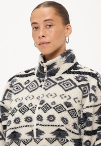 Pull en polaire crème avec un motif géométrique noir ; il présente un col montant et une fermeture éclair mi-longue. Tissu doux et texturé.