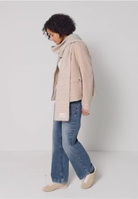 Chaquetón beige claro con dos bolsillos frontales, combinado con una bufanda de punto color crema. Unos jeans azules holgados y zapatos claros completan el atuendo. El modelo está de lado.