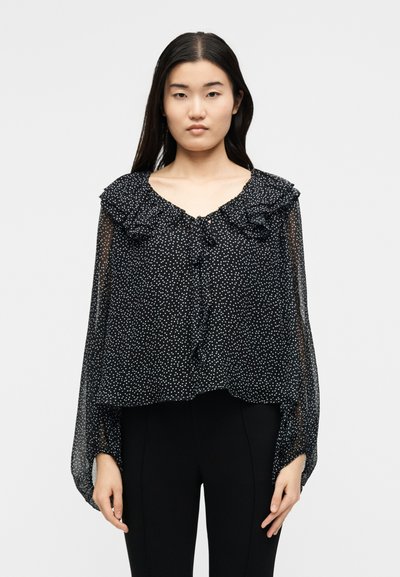 Γυναίκα που φοράει διάφανη μαύρη μπλούζα με λευκά πουά, με βολάν στον γιακά και μακριά μανίκια, συνδυασμένη με μαύρο παντελόνι, στέκεται μπροστά σε λευκό φόντο.