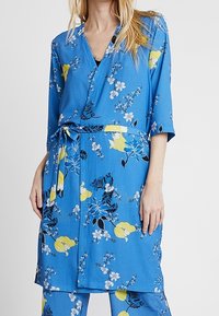 Modré zavinovací květinové šaty s krátkými rukávy, zdobené složitými žlutými a bílými květinovými vzory, s páskem v pase a měkkou texturou.