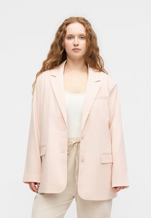 Νέα γυναίκα με μακριά, σγουρά κόκκινα μαλλιά, φοράει αέρινο, ανοιχτό ροζ σακάκι, λευκό τοπ και μπεζ παντελόνια με κορδόνι, μπροστά σε απλό φόντο.