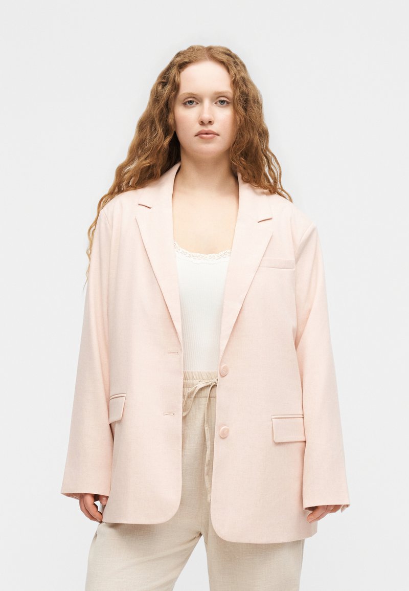 Νέα γυναίκα με μακριά, σγουρά κόκκινα μαλλιά, φοράει αέρινο, ανοιχτό ροζ σακάκι, λευκό τοπ και μπεζ παντελόνια με κορδόνι, μπροστά σε απλό φόντο.