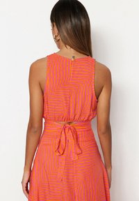 Robe sans manches, rose vif, avec un motif en zigzag rose plus foncé. Taille nouée et fermeture au dos avec un œillet. Texture lisse.