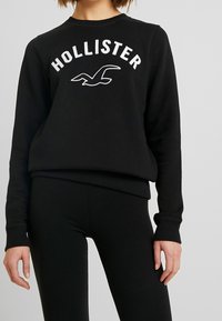 Černá mikina s kulatým výstřihem, žebrovanými manžetami a spodním lemem, s nápisem "HOLLISTER" bílými velkými písmeny a logem racka.