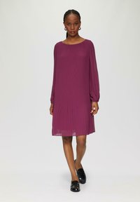 Robe plissée en prune profond, avec des manches longues et un col arrondi. Tissu doux qui tombe délicatement jusqu'à mi-thigh, associée à des chaussures noires.
