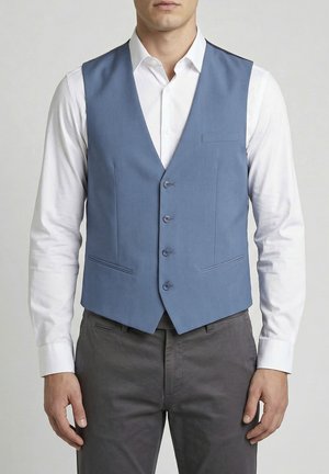 Uomo che indossa un gilet azzurro chiuso da bottoni sopra una camicia bianca e pantaloni grigio scuro, in piedi con le mani rilassate ai lati.