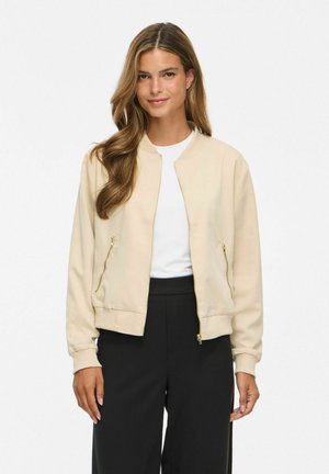 Femme aux longs cheveux bruns portant une veste beige zippée sur une chemise blanche et un pantalon noir, debout devant un fond blanc uni.