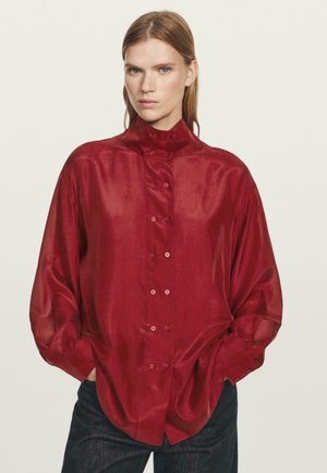 Blusa di seta rossa con colletto alto, frontale con bottoni e maniche lunghe e morbide. Tessuto liscio con una leggera lucentezza, con bottoni tonali.