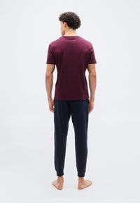 Άνδρας με σγουρά μαλλιά στέκεται ξυπόλυτος, γυρισμένος πλάτη, φοράει μπορντό μπλουζάκι και μπλε παντελόνι σε απλό λευκό φόντο.