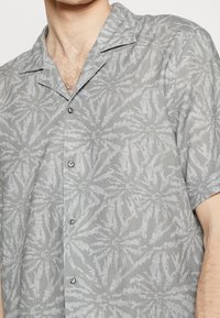 Szara koszula z krótkim rękawem z wzorem liści, zapinana na guziki z przodu, z kołnierzem typu kent, wykonana z fakturowanej tkaniny.