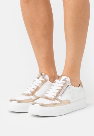 Zapatillas blancas con detalles metálicos dorados, materiales de cuero y sintéticos, diseño con cordones, cuello acolchado y detalle de cremallera lateral.