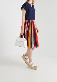 Femme portant un chemisier bleu marine et une jupe plissée à rayures colorées, tenant un sac à main blanc et portant des sandales compensées blanches.