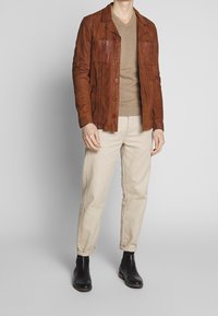 Brun läderjacka med knäppning framtill och två bröstfickor, bärs över en beige V-ringad tröja och matchas med ljusbeiga byxor.