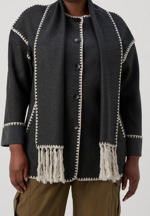 Person trägt eine dunkelgraue Jacke mit weißen Zickzack-Borten und passenden Schal mit Fransen, kombiniert mit olivgrüner Hose.