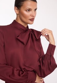 Burgundowa bluzka z wiązanym kokardą przy kołnierzy, rękawami w stylu puff oraz guzikami na mankietach, wykonana z gładkiego materiału.