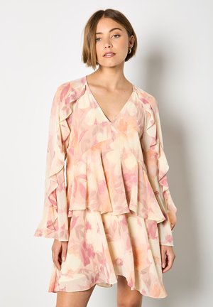 Giovane donna che indossa un vestito a fiori pastello con scollo a V, maniche lunghe con volant e gonna a strati, in piedi davanti a uno sfondo bianco uniforme.