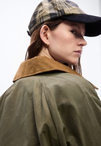 Γυναίκα που φοράει καρό καπέλο, ελιά πράσινο μπουφάν με καφέ γιακά από βελούδο, και μικρό χρυσό σκουλαρίκι σε σχήμα κρίκου, κοιτάζοντας πάνω από τον ώμο της.