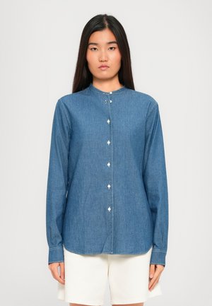 Femeie îmbrăcată cu o cămașă albastră din denim cu mânecă lungă, prevăzută cu butoane albe, și pantaloni scurți albi, în fața unui fundal simplu.