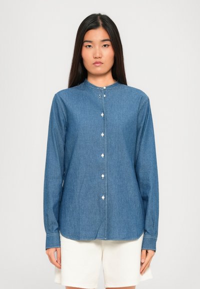 Femme portant une chemise en denim bleu à manches longues avec des boutons blancs et un short blanc devant un fond uni.