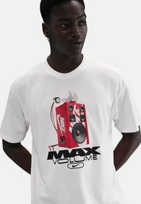 Hvid bomulds T-shirt med et rødt grafik af en boombox, med teksten "MAX VOLUME" og Nike-logoet i sort. Enkel crew neck design.