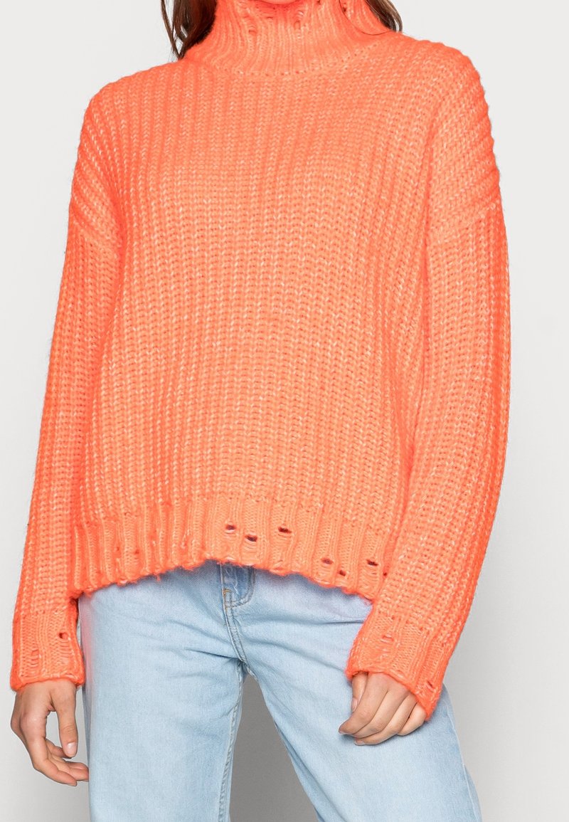 Pull à col roulé en gros tricot de couleur corail vif. Présente un ourlet et des manches côtelés avec de petits trous. Associé à un jean bleu clair.