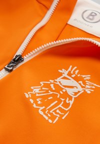 Orange syntetisk stof med en hvid lynlås og et fremtrædende hvidt grafisk design. Mærket har et rundt logo med "B" og "BOGNER."