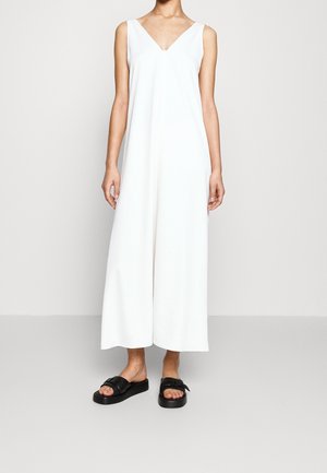 Personne portant une combinaison blanche sans manches à jambes larges et des sandales noires sans bride, debout sur un fond uni.