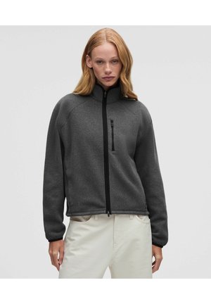 Mujer con cabello liso castaño claro lleva chaqueta de forro polar gris oscuro con cremallera y pantalones beige claro, de pie frente a un fondo claro y liso.