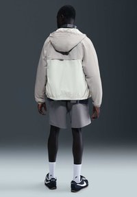 Hombre de espaldas con chaqueta con capucha de dos tonos, pantalones cortos grises, calcetines blancos y zapatillas negras Nike sobre un fondo gris liso.