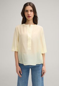 Blusa giallo chiaro, semi-trasparente, con scollatura rotonda, maniche corte e una patta centrale con piccoli bottoni; abbinata a jeans blu.