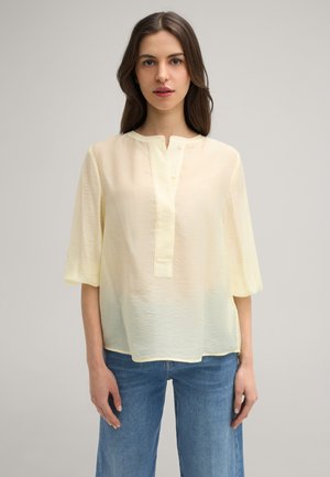 Blusa giallo chiaro, semi-trasparente, con scollatura rotonda, maniche corte e una patta centrale con piccoli bottoni; abbinata a jeans blu.