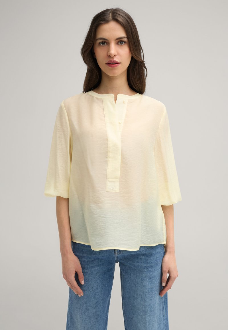Blusa giallo chiaro, semi-trasparente, con scollatura rotonda, maniche corte e una patta centrale con piccoli bottoni; abbinata a jeans blu.