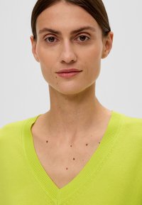 Sweter w serek w jasnym limonkowym kolorze, z prążkowanymi wykończeniami i miękką, cienką dzianiną. Widoczne niedoskonałości skóry na szyi.