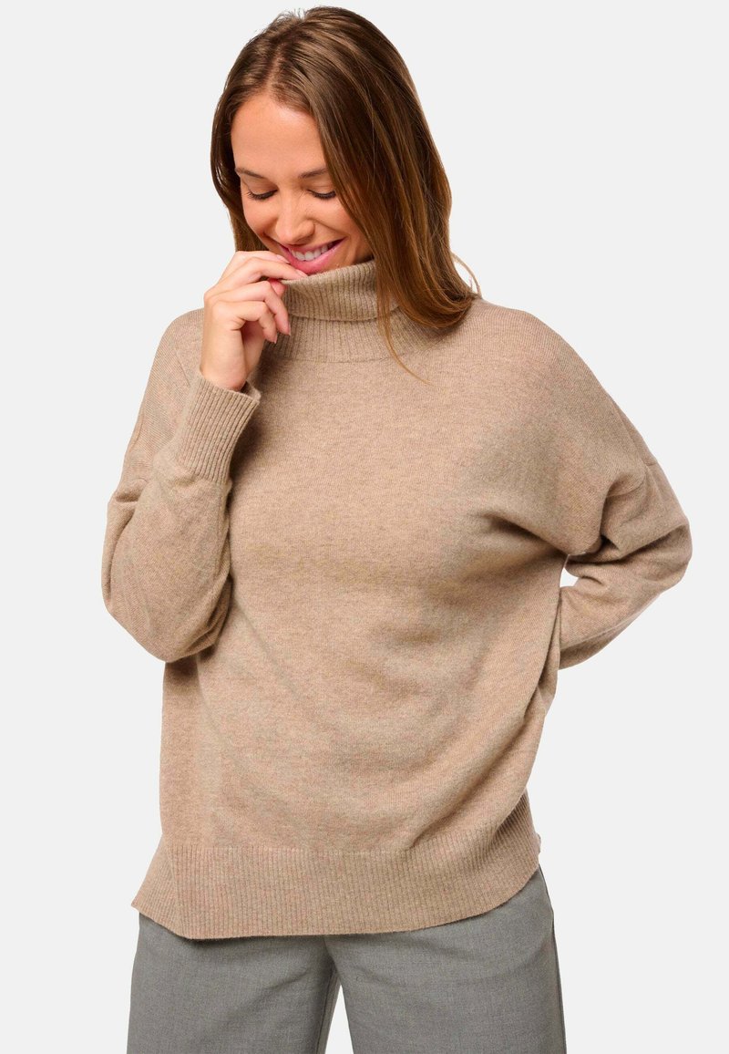 Pull en col roulé beige avec une coupe ample, des poignets et un ourlet côtelés. Texture douce avec des variations de couleur subtiles. Associé à un pantalon gris.