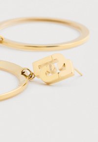 Guldtonede metal hoops øreringe med et chunky kædested, der forbinder posten og hoop'en på en hvid baggrund.