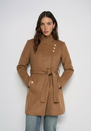 Femme portant un manteau en laine camel ceinturé avec des boutons dorés, debout devant un fond clair uni.