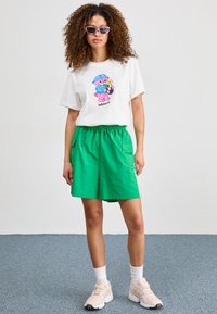 Weißes T-Shirt mit buntem Bär-Print, kombiniert mit grünen Shorts mit seitlichen Taschen. Das Outfit wird durch hellrosa Sneaker und Sonnenbrille abgerundet.