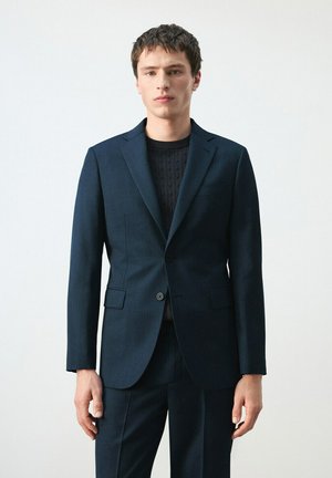 Jonge man draagt een donkerblauw op maat gemaakt colbert en bijpassende broek over een zwarte, gestructureerde trui tegen een eenvoudige lichte achtergrond.