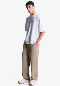 Tricou oversized din bumbac de culoare gri deschi și pantaloni de trening bej. Pantalonii au buzunare laterale. Modelul poartă adidași albi cu accente bej.