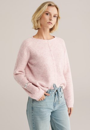 Lichtroze, geribbelde gebreide trui met een ronde hals en knoopsluiting aan de voorkant. Gecombineerd met hoge, lichtgewade jeans.