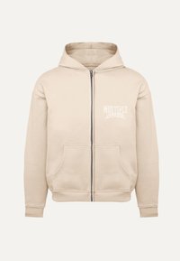 Sudadera con cremallera beige, hecha de una tela suave, con capucha ajustable, bolsillos frontales y un pequeño logo blanco en la parte superior izquierda del pecho.
